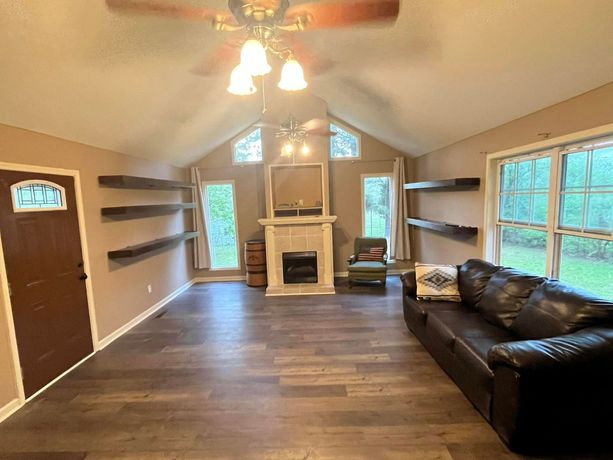 Empty living room with vaulted ceiling, fireplace, built-in shelves, and a dark leather sofa by large windows