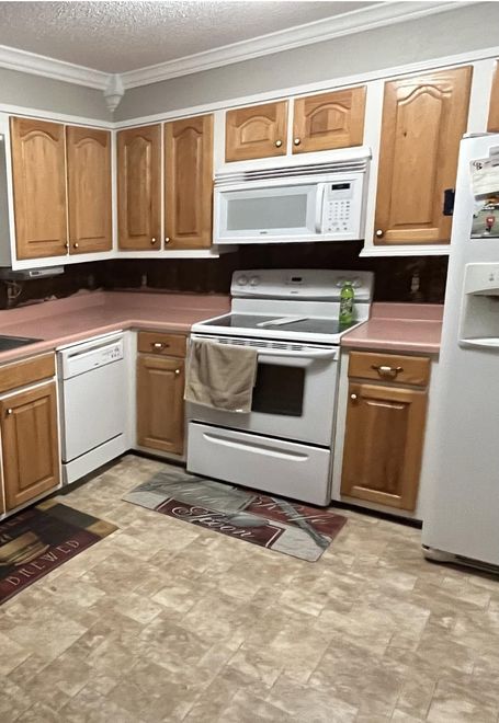 Small kitchen with wood cabinets, white appliances, and a beige patterned floor rug.