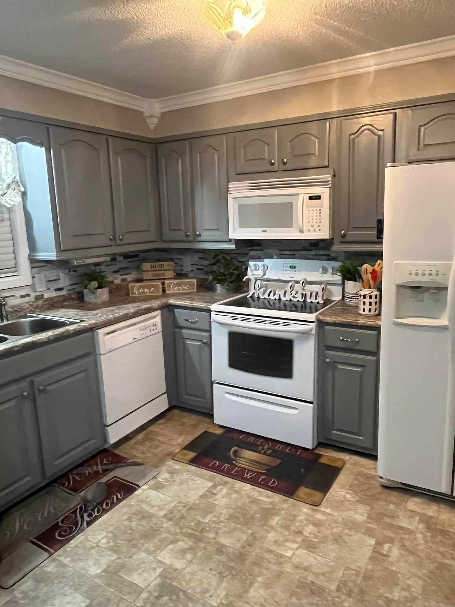 Small kitchen with gray cabinets, white appliances, beige rug, and window curtains