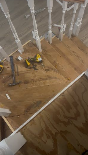 Two yellow power tools on a wooden floor beneath white stair railings, with a plywood panel in the foreground