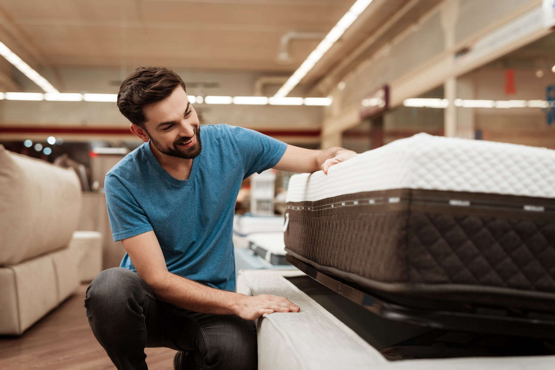 View of a neutral bed with a soft and white mattress set and pillows. A young bearded man is testing a mattress set at a furniture store.