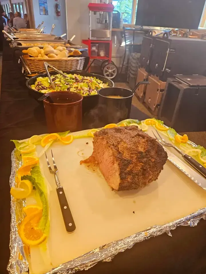 Buffet tray with a roasted meat centerpiece and serving utensils, surrounded by vegetables in a kitchen setting