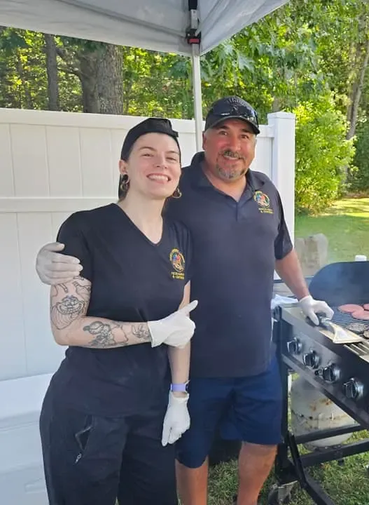 Two volunteers in navy shirts and gloves smile beside a grill under a canopy outdoors.