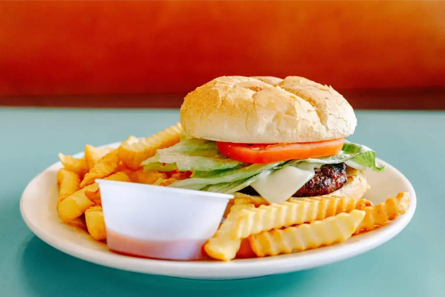 Burger with crinkle-cut fries and a dipping sauce on a white plate against a teal table.