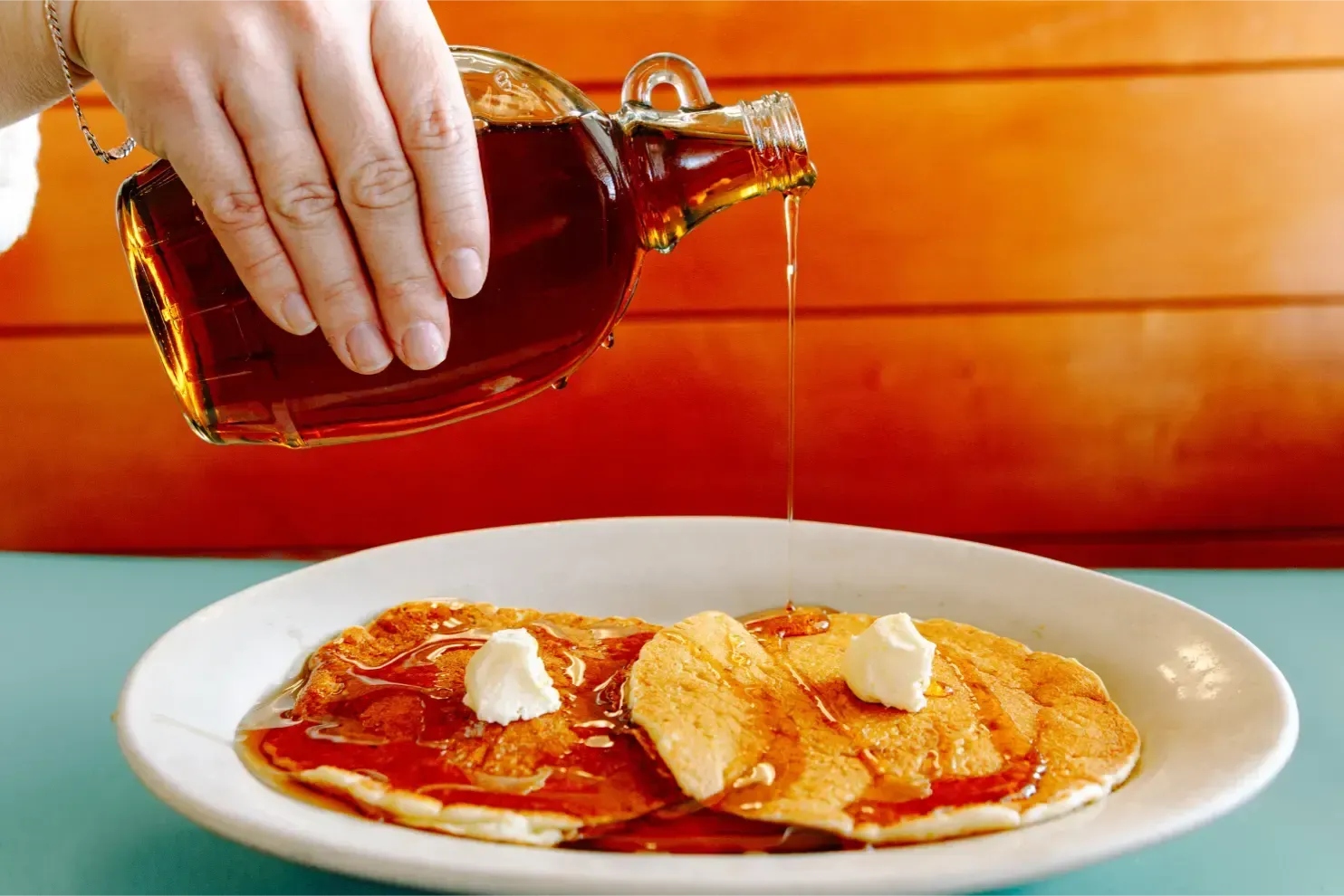Hand pouring syrup over a plate of pancakes with butter on a white dish