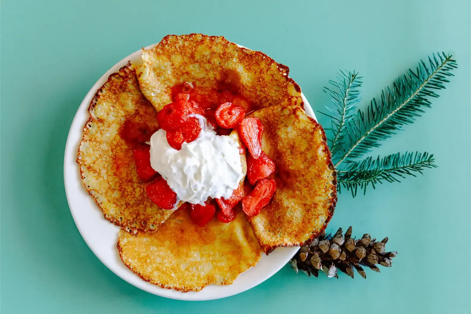 Pancakes topped with whipped cream and strawberries on a white plate with a pine sprig on a teal background