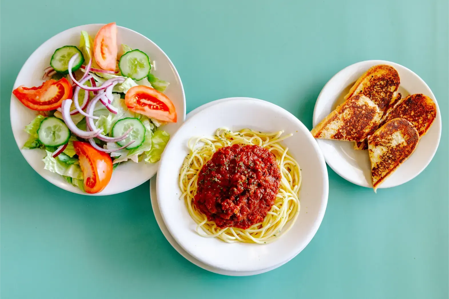Three plates of food on a mint-green table: salad, spaghetti with red sauce, and toasted bread