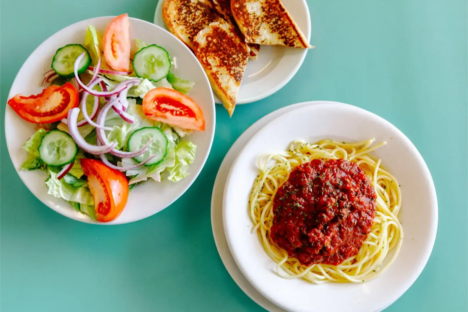 Three plates of food on a teal table: spaghetti, salad, and toasted bread slices.