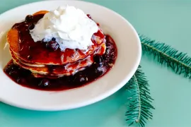Stack of pancakes topped with whipped cream and berry sauce on a white plate, on a turquoise table