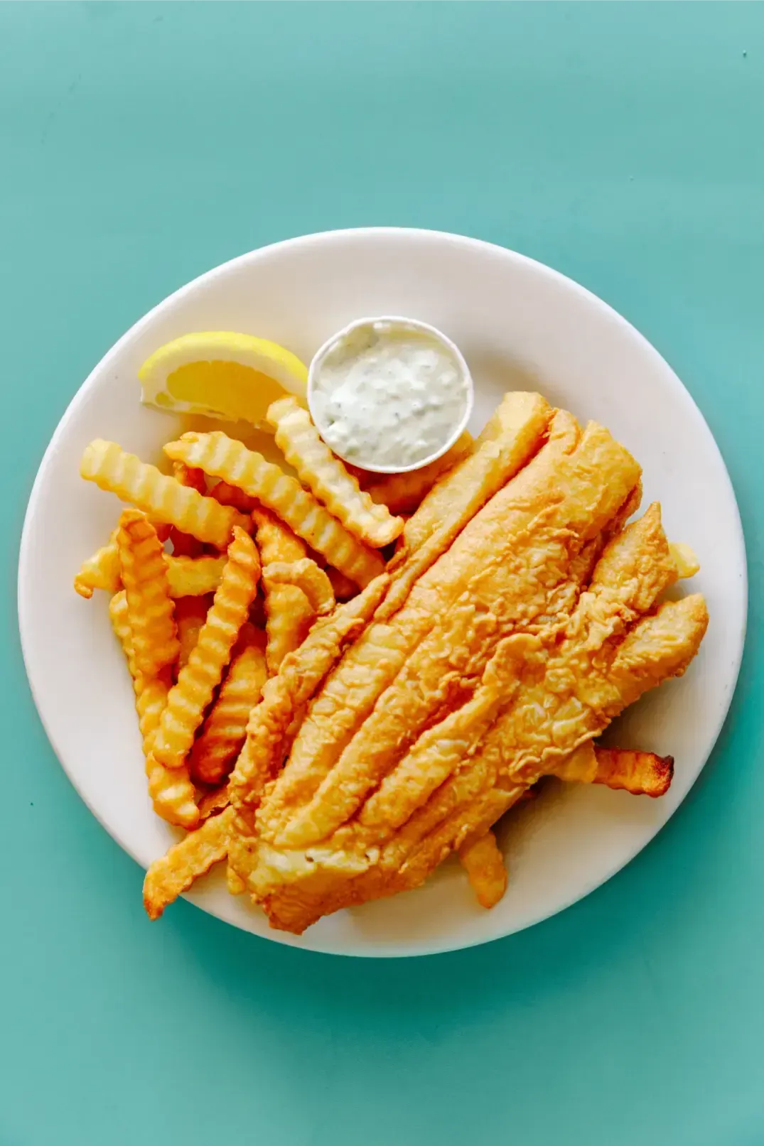 Plate of fried fish and crinkle-cut fries with lemon and tartar sauce on a teal background
