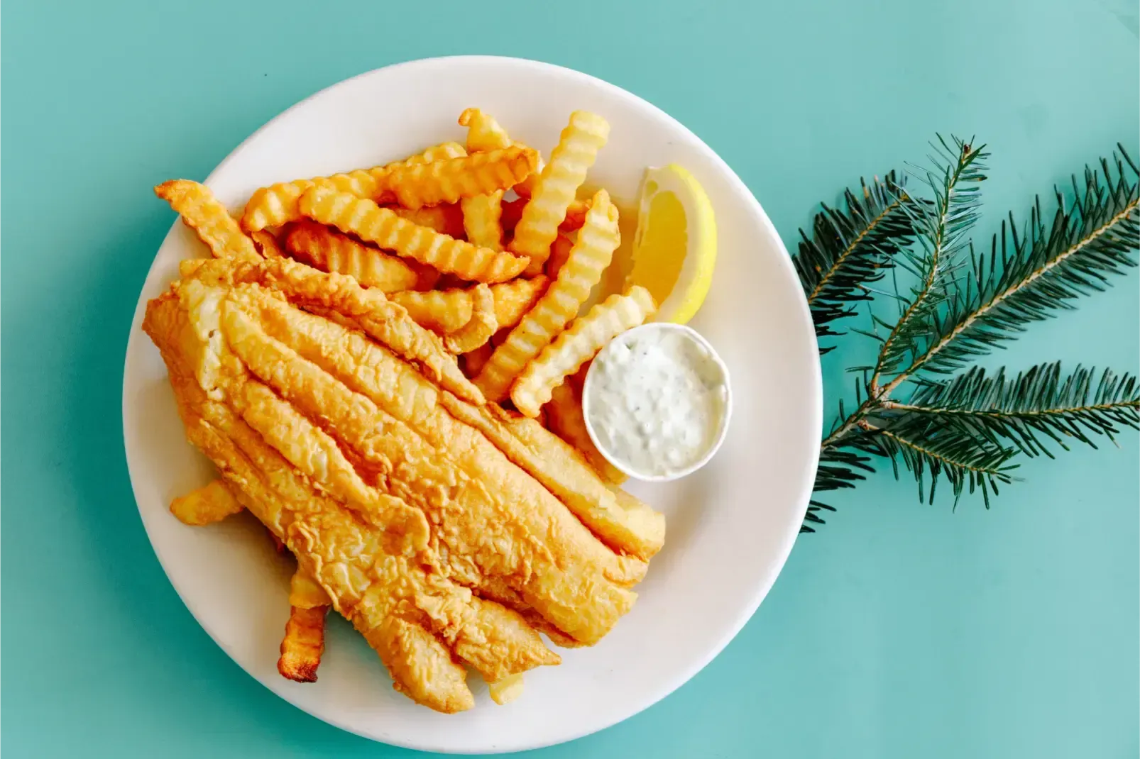 Fried fish, crinkle fries, and tartar sauce on a white plate with greenery on a teal background