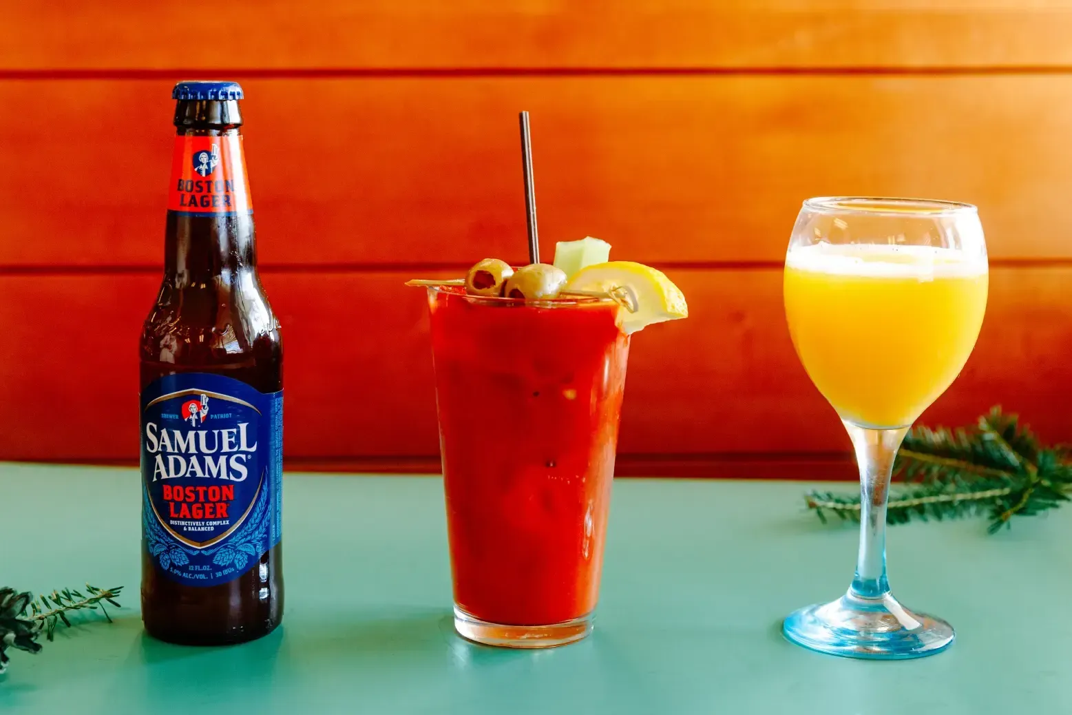 Samuel Adams beer bottle beside a red cocktail and an orange drink on a teal table