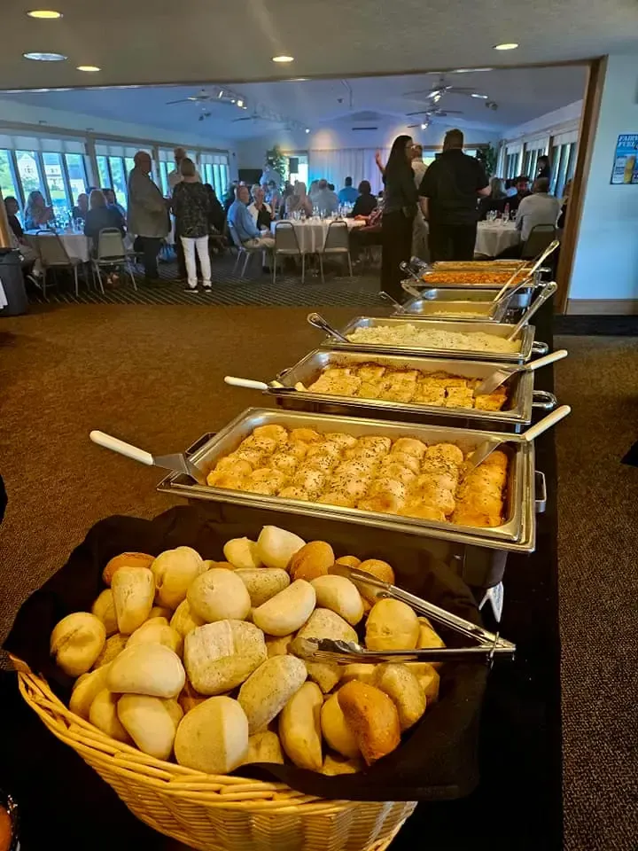 Buffet table with bread rolls and trays of hot dishes in a bright dining room with people in the background