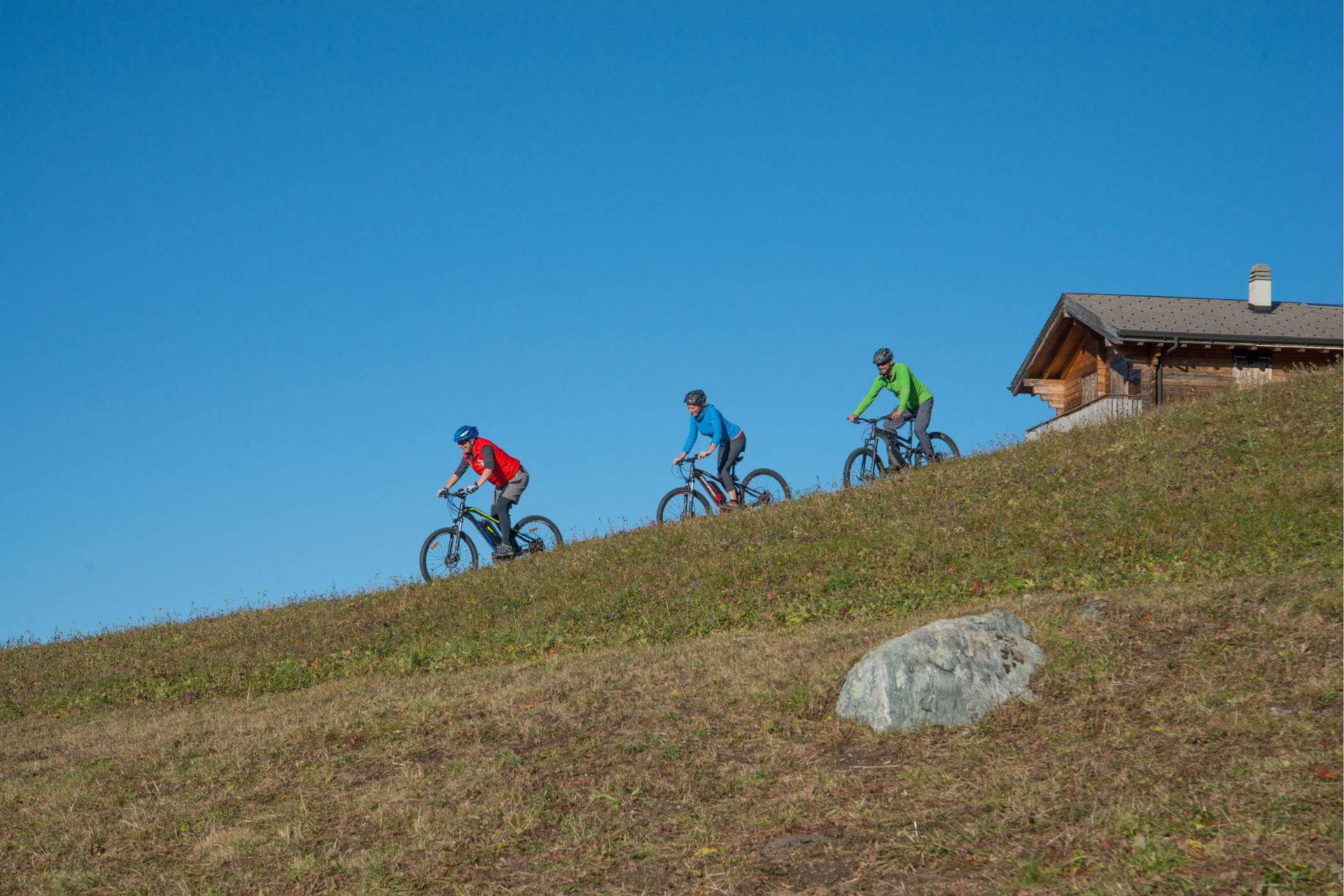 Trois cyclistes montent à vélo une colline herbeuse par une journée ensoleillée, près d'une cabane en bois.