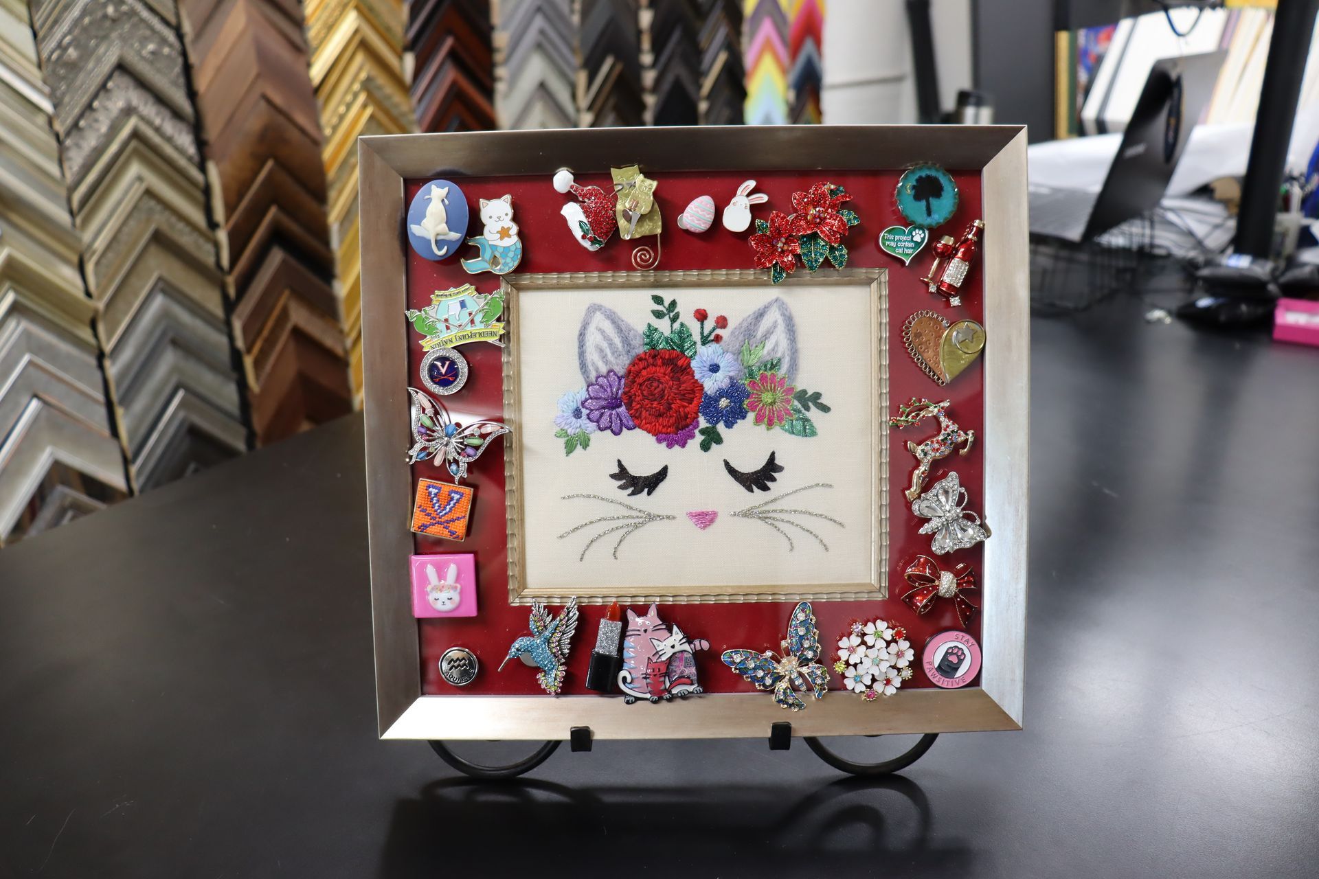 An embroidered cat face with a floral crown, framed and surrounded by various colorful enamel pins on a red mat.