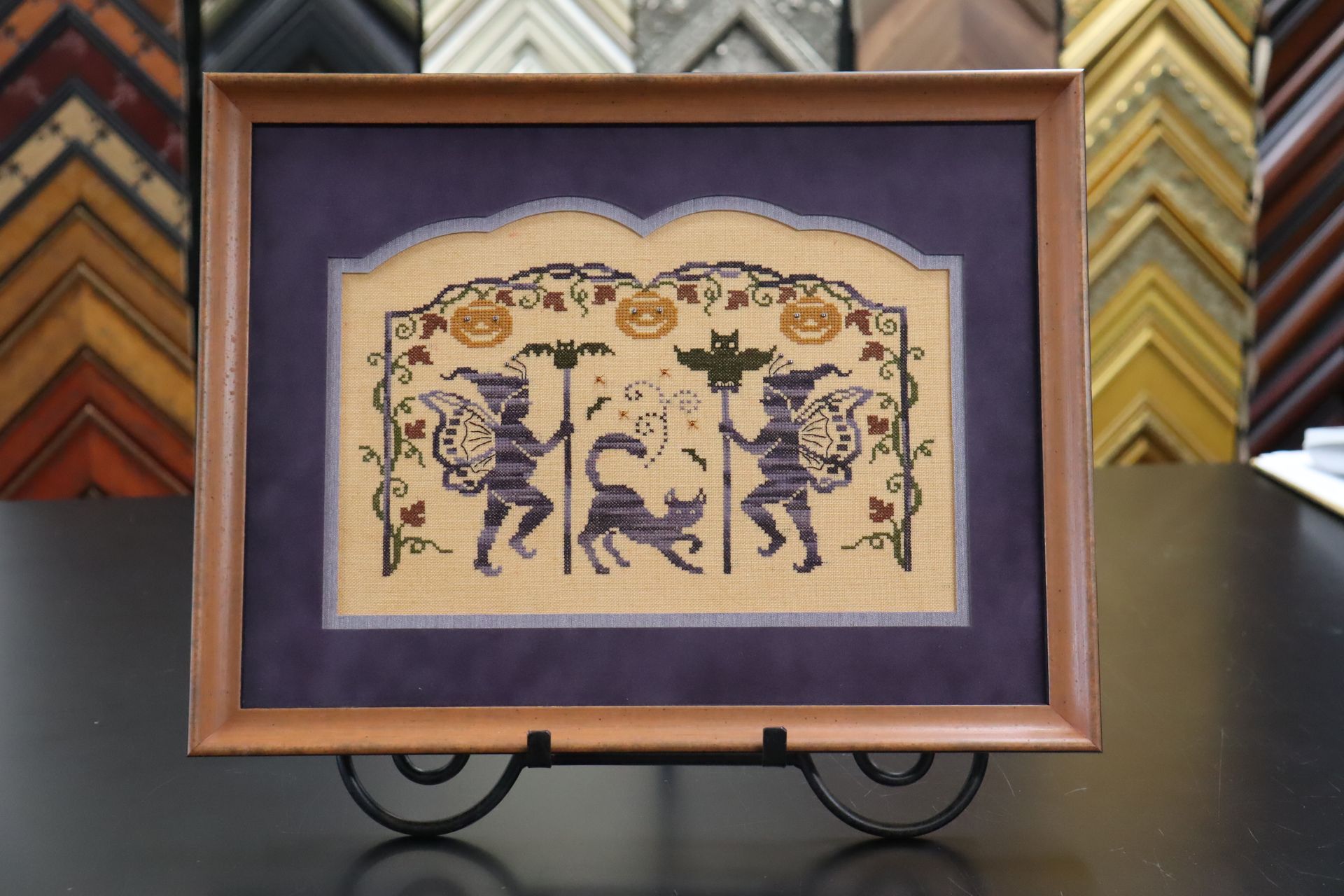 A framed cross-stitch artwork featuring two figures and a cat, decorated with pumpkins and vines on a tan background.
