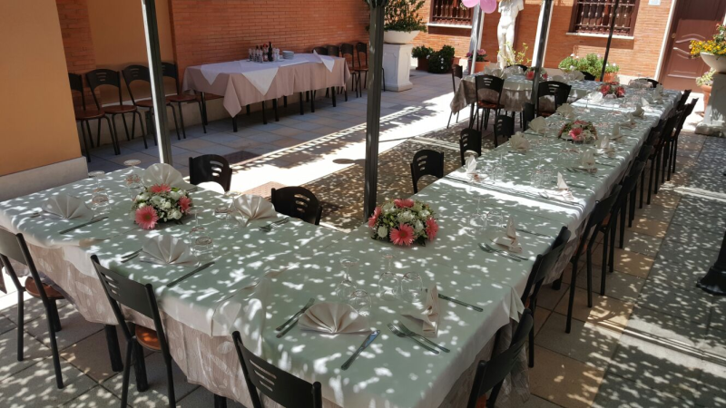 Lunghi tavoli da banchetto con tovaglie a motivi verdi e bianchi, centrotavola di fiori rossi e sedie nere in un cortile.