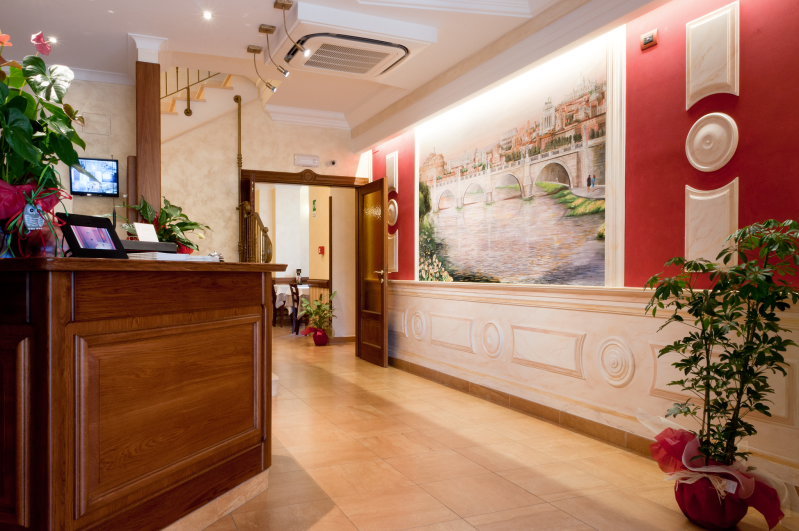 Elegante hall d'albergo con bancone della reception in legno, pareti rosa e grandi quadri floreali.