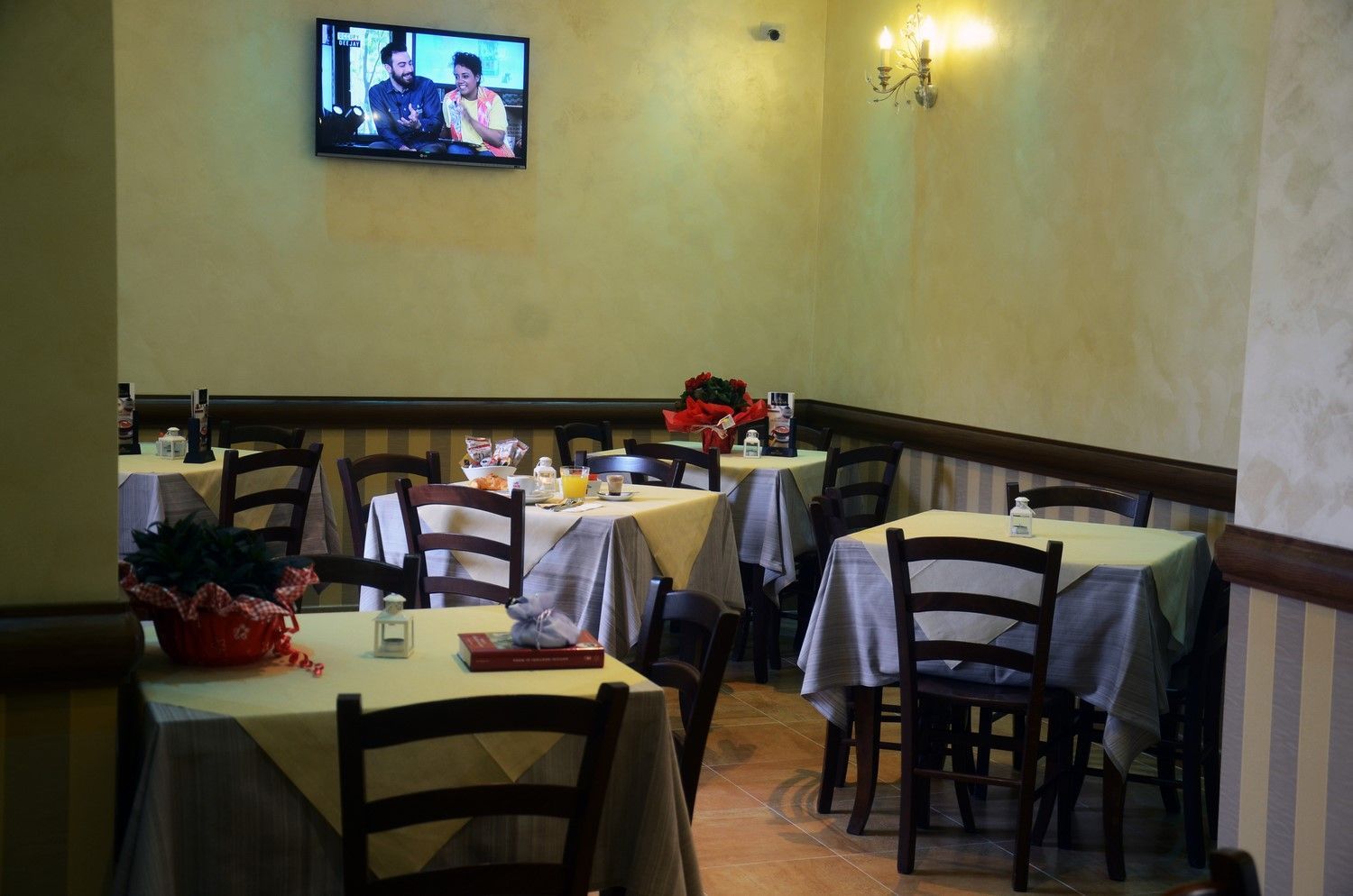Piccola sala da pranzo di un ristorante con tavoli vuoti, tovaglie bianche, sedie e un televisore a parete.