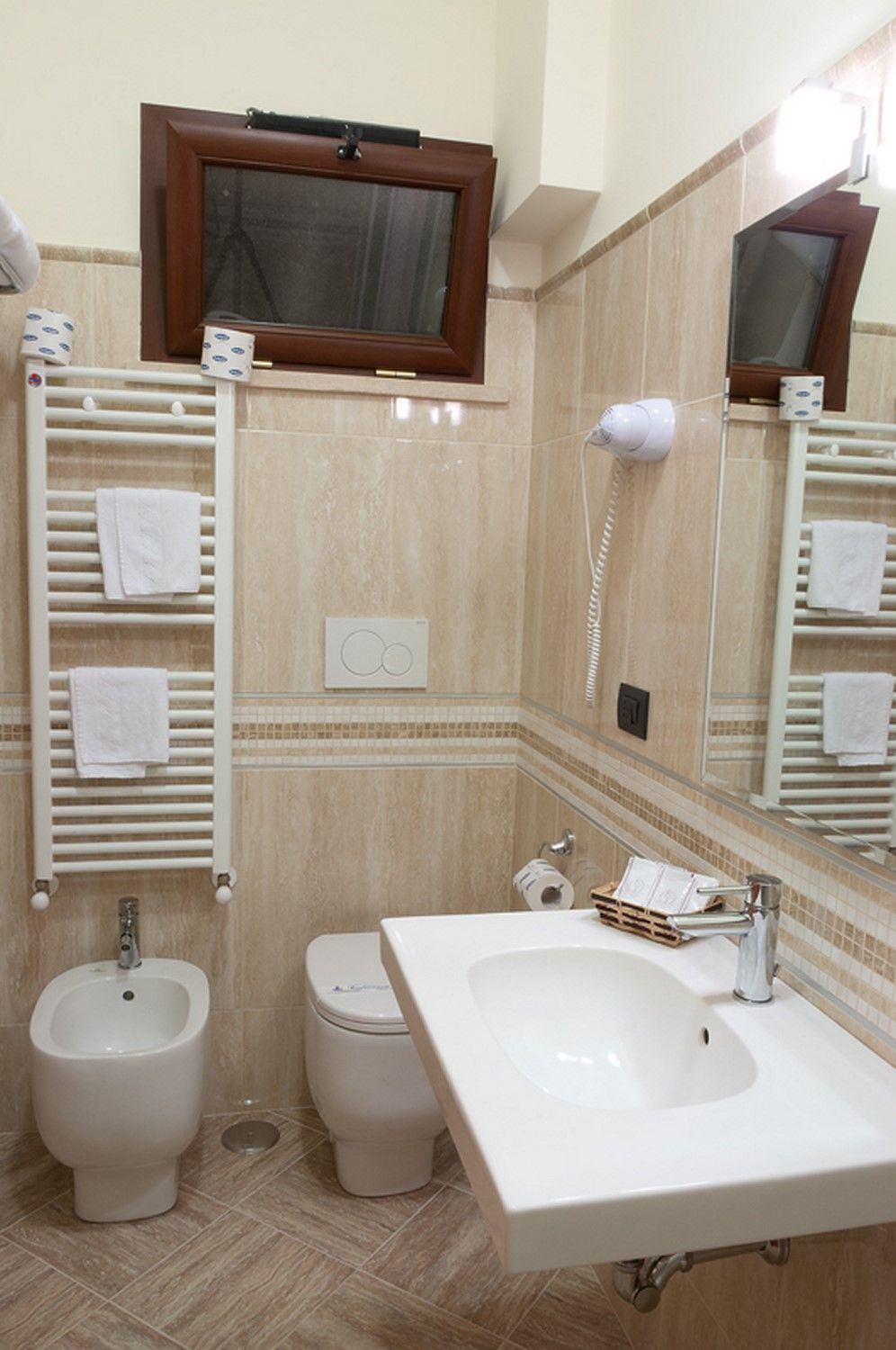 Piccolo bagno beige con WC, lavabo, bidet, specchio e termoarredo sotto una finestra.
