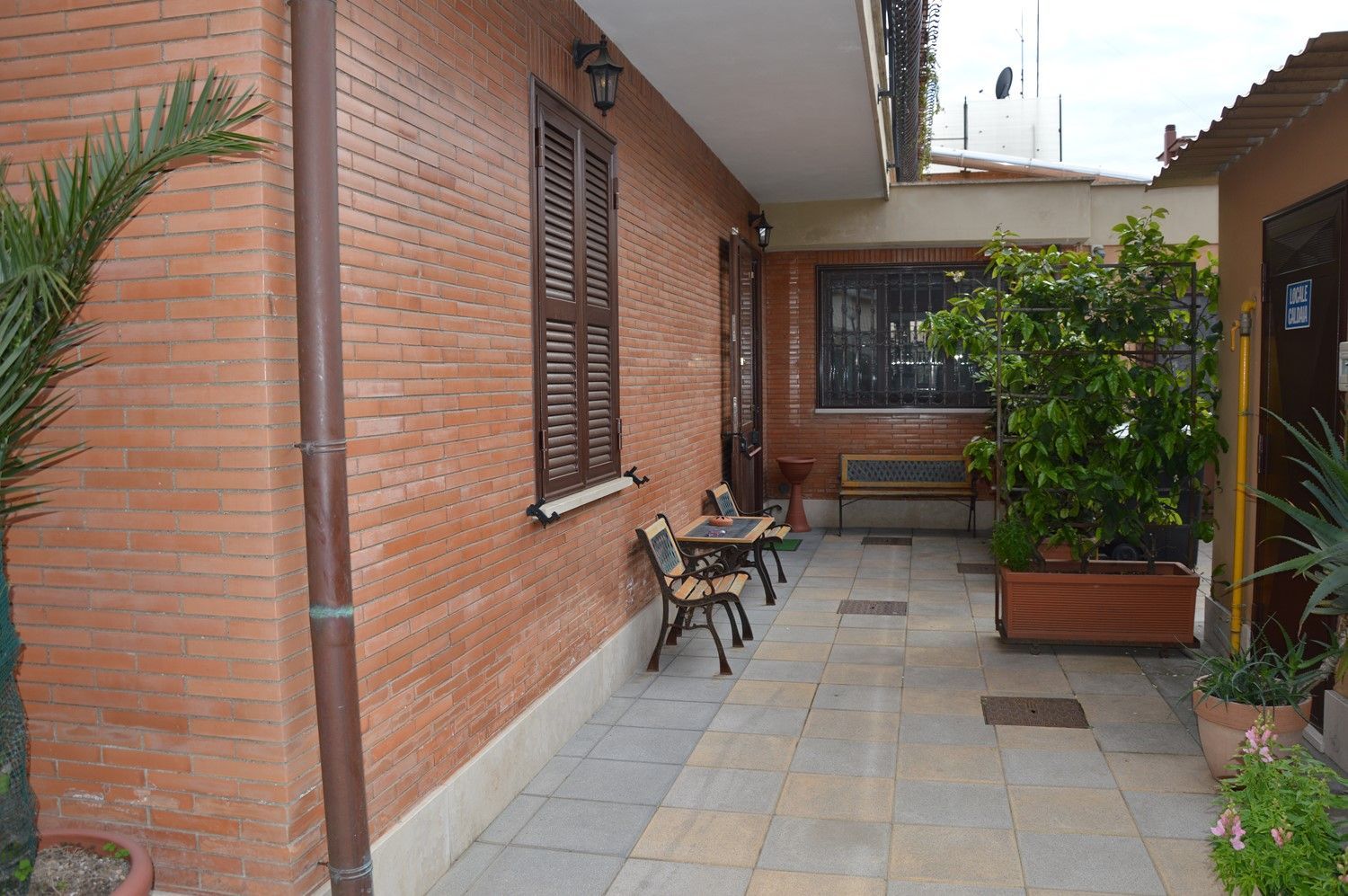 Stretto passaggio pedonale in mattoni nel cortile interno, con piante in vaso, sedie e finestre lungo il muro.