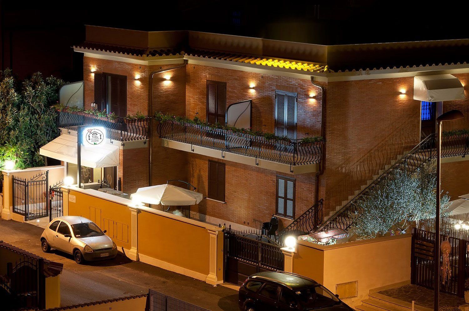 Vista notturna di un edificio in mattoni con balconi illuminati, un'auto bianca parcheggiata all'esterno e un muro con cancello.