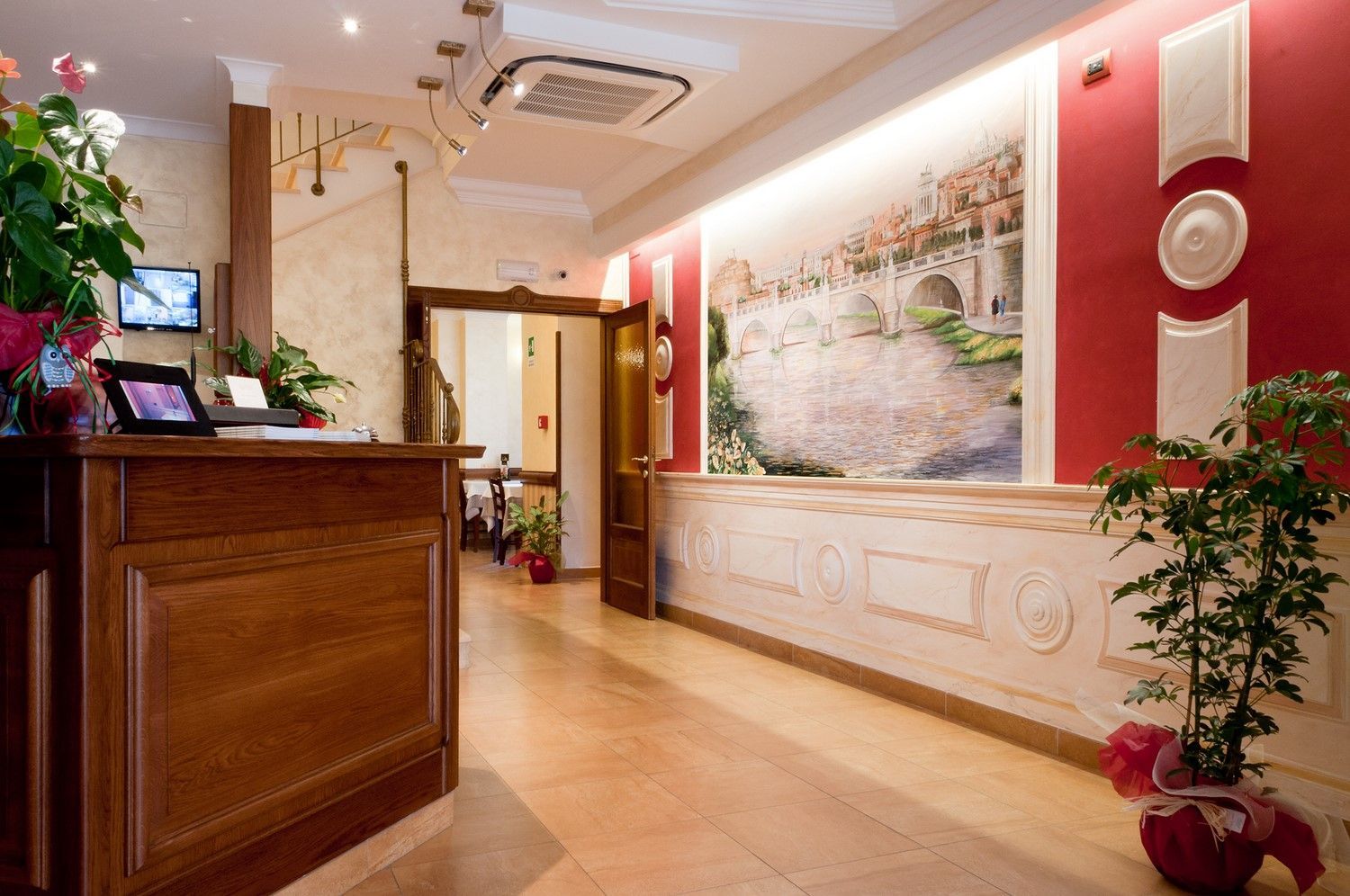 Hall dell'hotel con bancone della reception in legno, pareti rosse, piante in vaso e un grande murale paesaggistico.