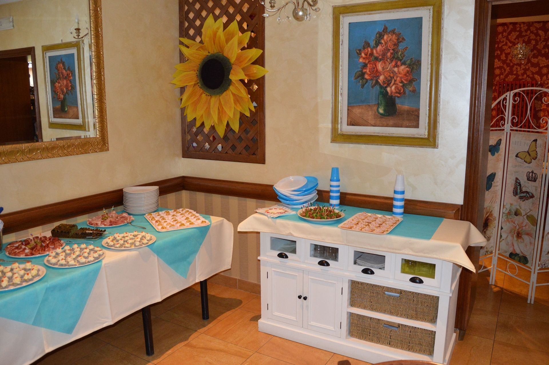Sala da pranzo luminosa con tavoli apparecchiati per un pasto, quadri con girasoli alle pareti e credenza bianca con decorazioni blu.