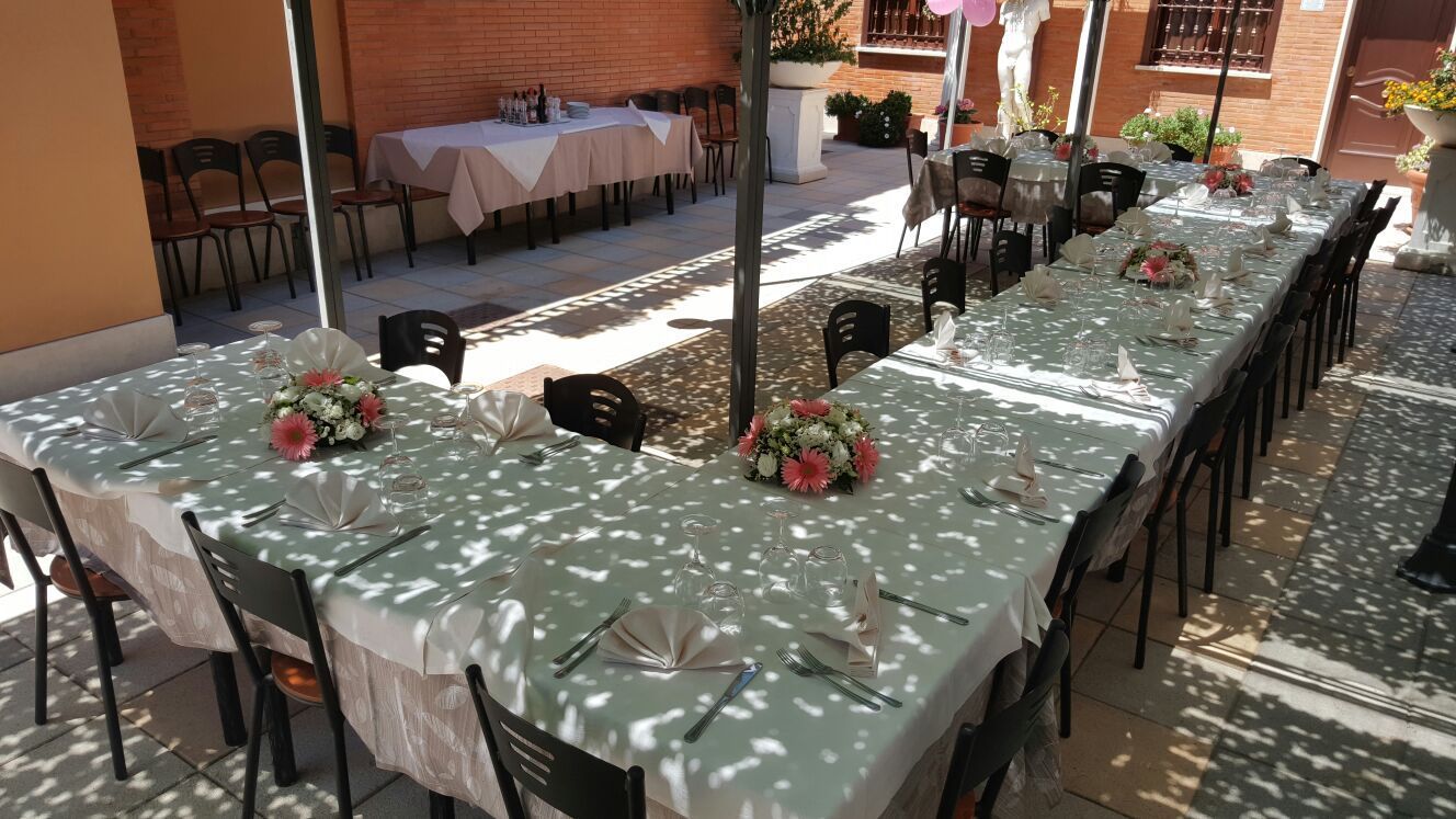 Lunghi tavoli da pranzo all'aperto con tovaglie bianche, sedie nere e centrotavola floreali rossi alla luce del sole.