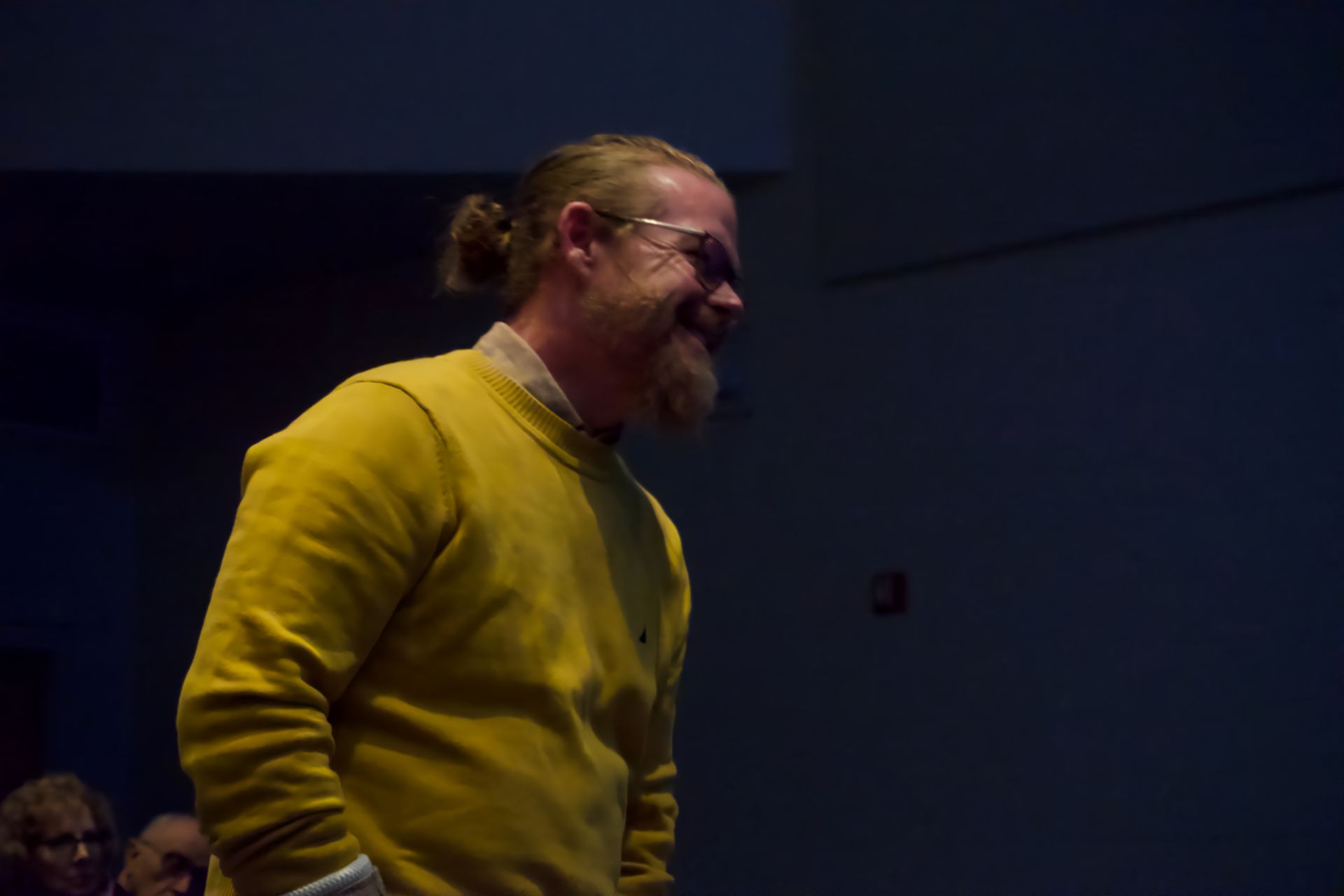 A man with a ponytail and glasses is wearing a yellow sweater.