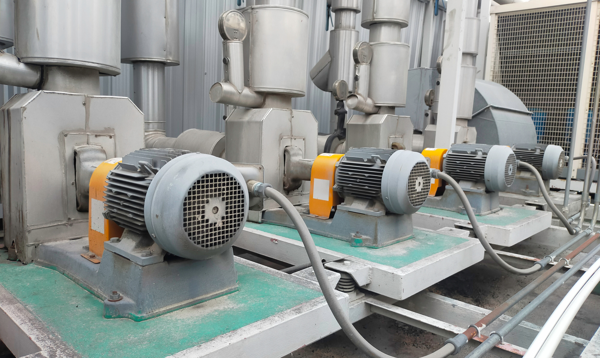Three industrial pumps with electric motors, metal and silver tones, on green platforms, with gray cabling. Three industrial pumps with electric motors, metal and silver tones, on green platforms, with gray cabling.