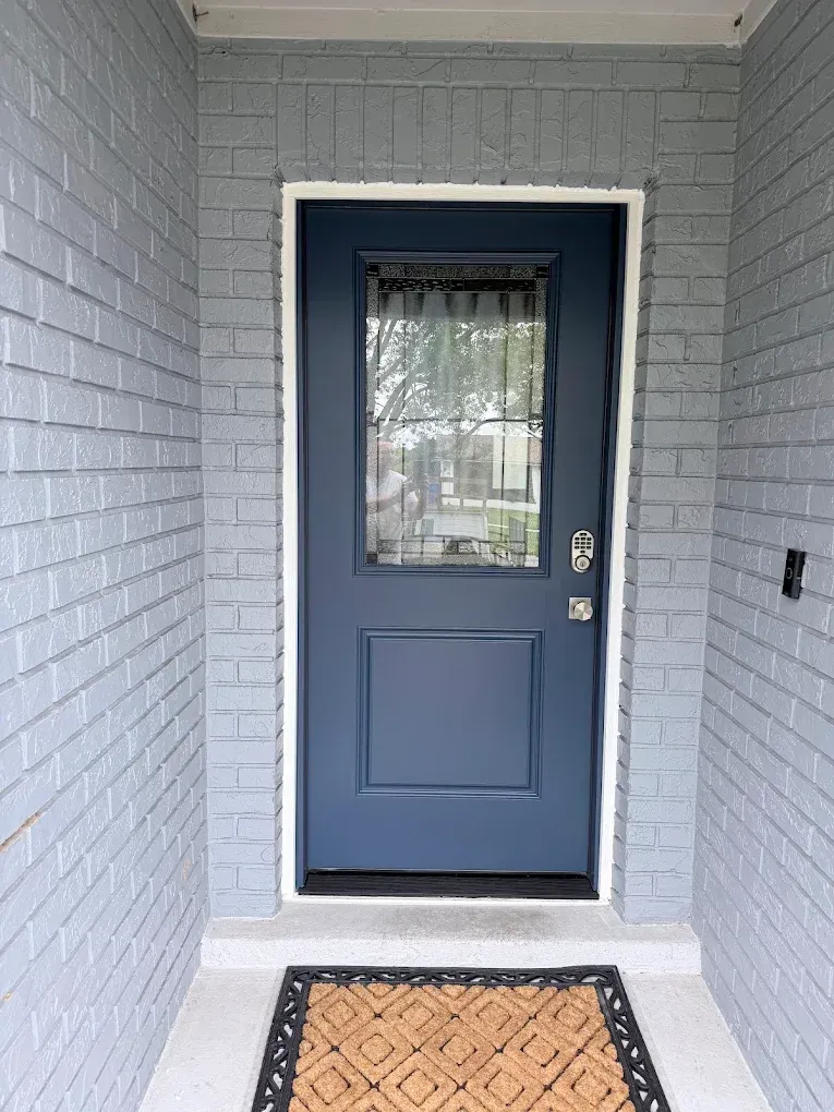 Front porch with a blue door, small window, gray brick walls, and a patterned doormat.