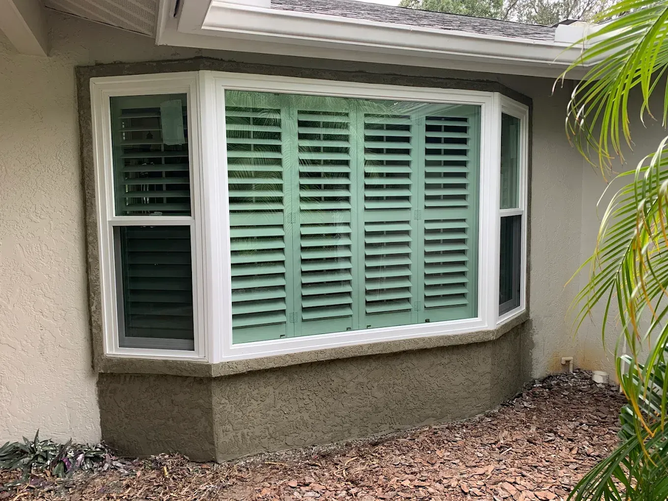 Bay window with mint-green shutters on a stucco house, bordered by white trim and leaf-covered ground