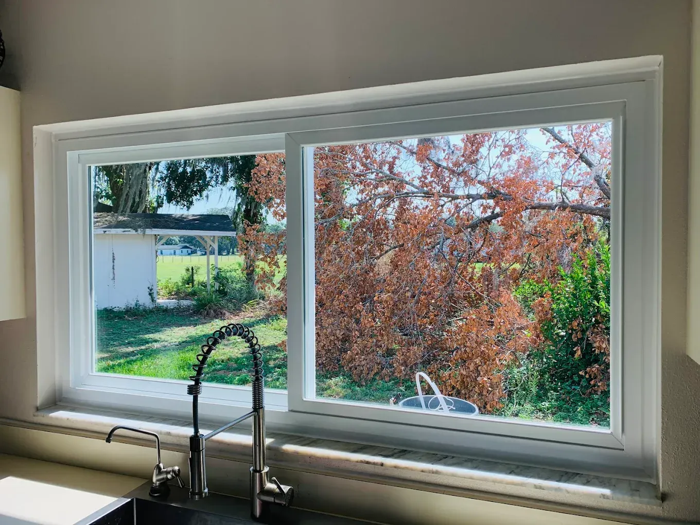 Kitchen window overlooking a backyard with green lawn, trees, and red autumn foliage.