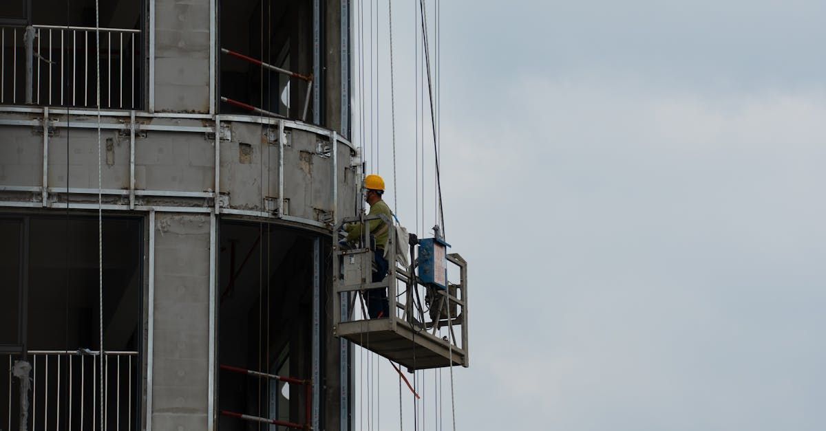 Worker on a suspended platform inspecting a tall industrial structure with cables and metal tanks