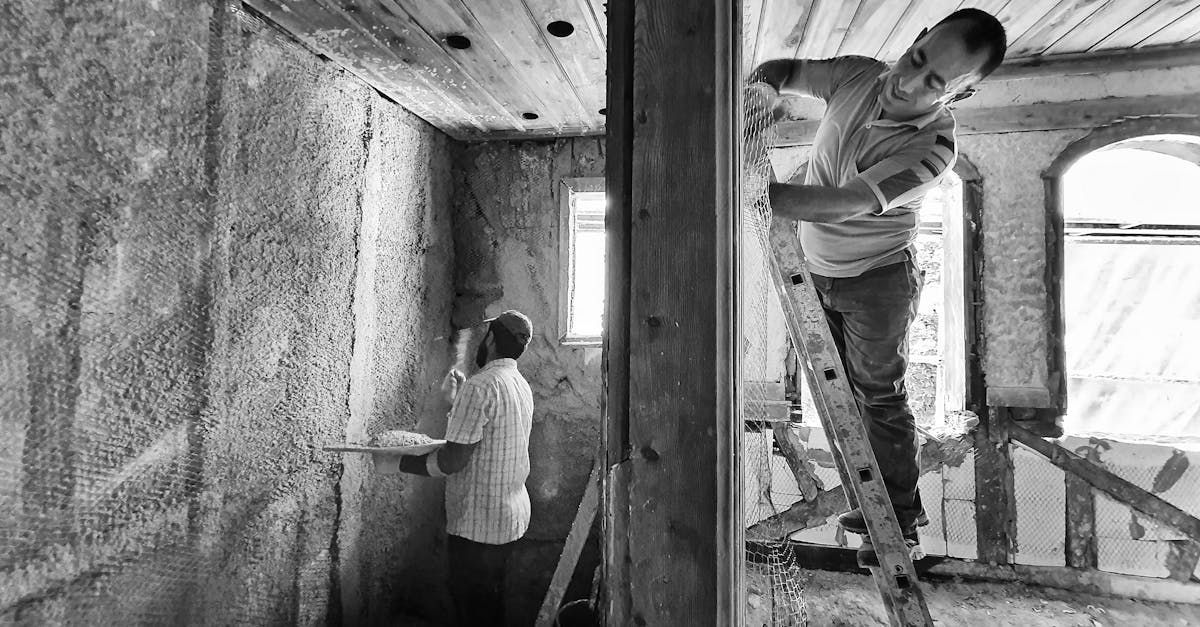 Two people working on a construction site, one inside near a window and one standing on scaffolding outside.