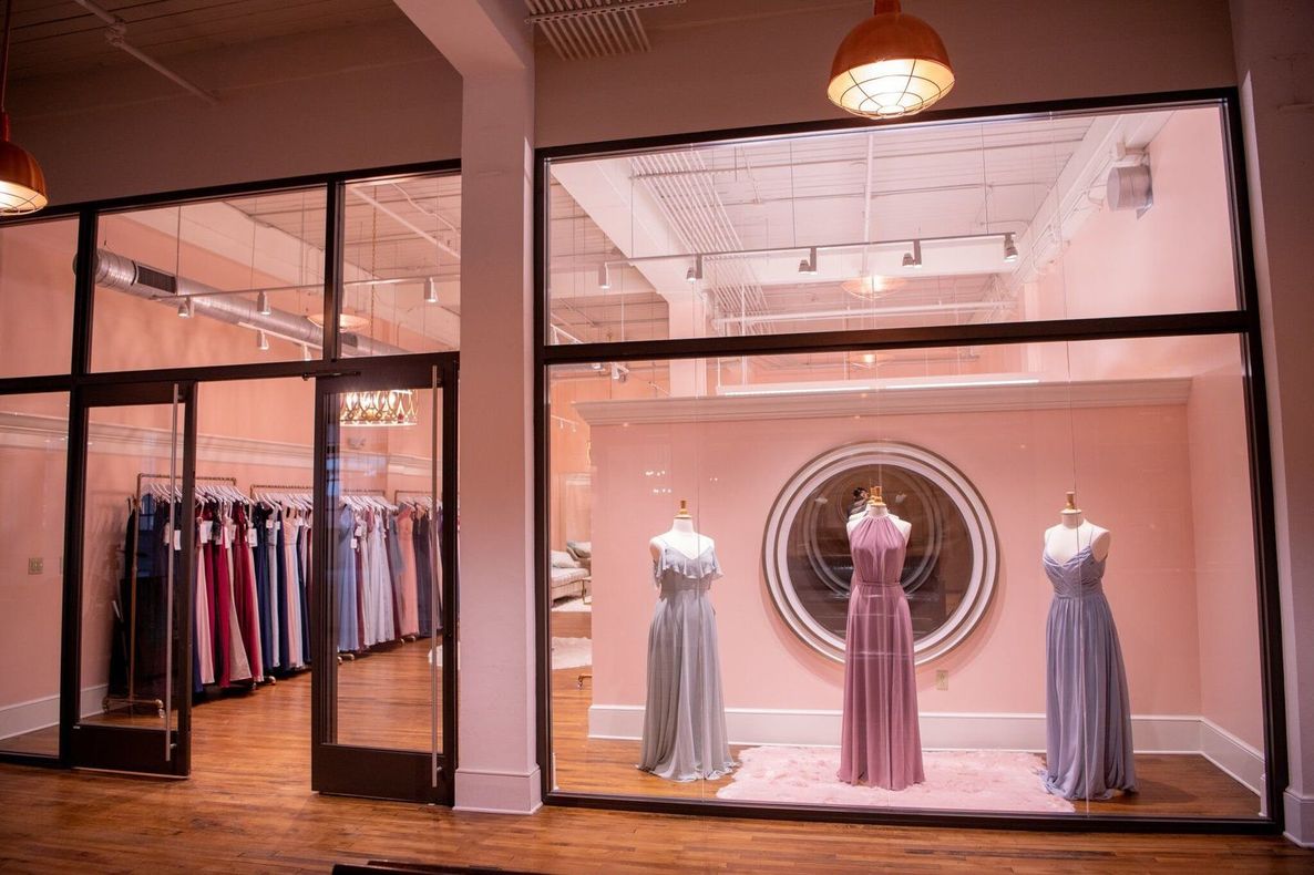 Boutique storefront with three dresses on mannequins and clothing racks inside, warmly lit at dusk