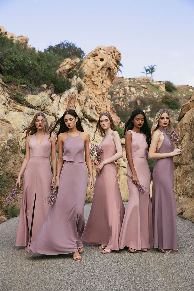 Five women in matching pink gowns walk outdoors on a gravel path among rocky hills.