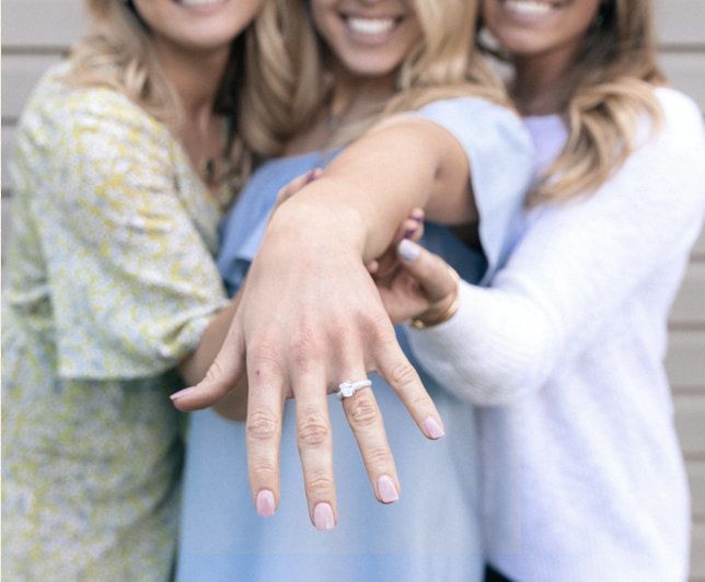 Three women smile outdoors, with a hand extended forward showing an engagement ring.