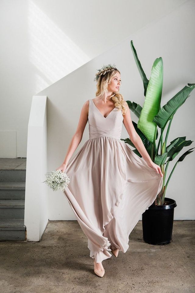 Woman in a blush dress holding flowers, standing by a large potted plant on indoor stairs.