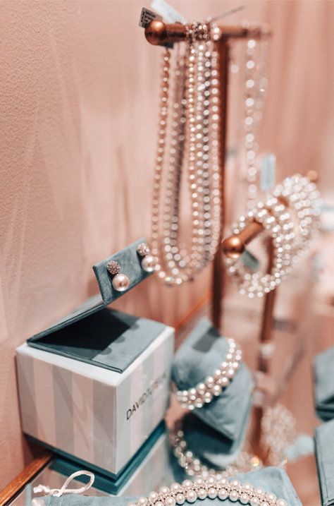 Pearl jewelry displayed on rose-gold stands beside an open ring box on a pink background