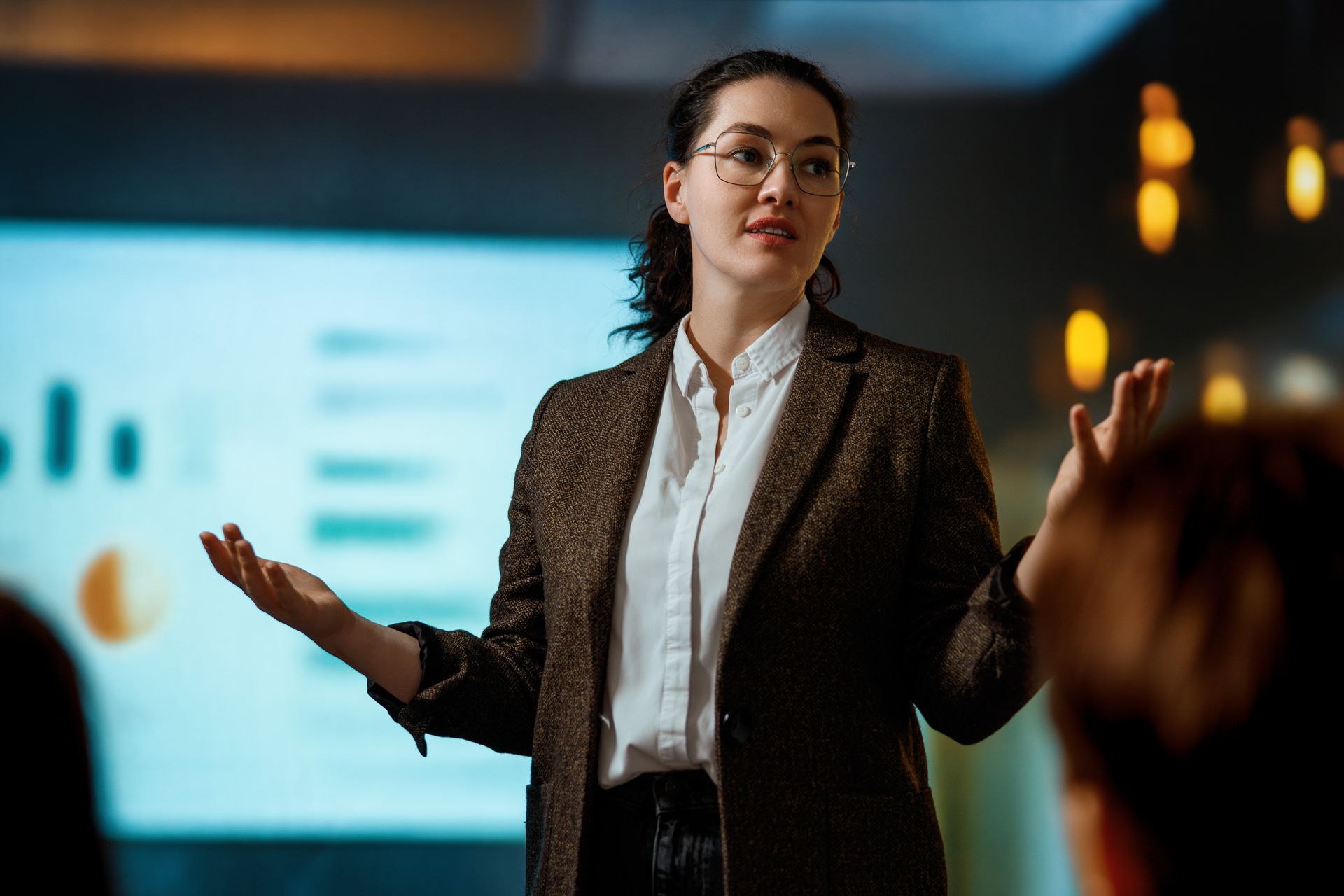 Presenter speaking in front of a projected slide, gesturing with both hands in a dim conference room