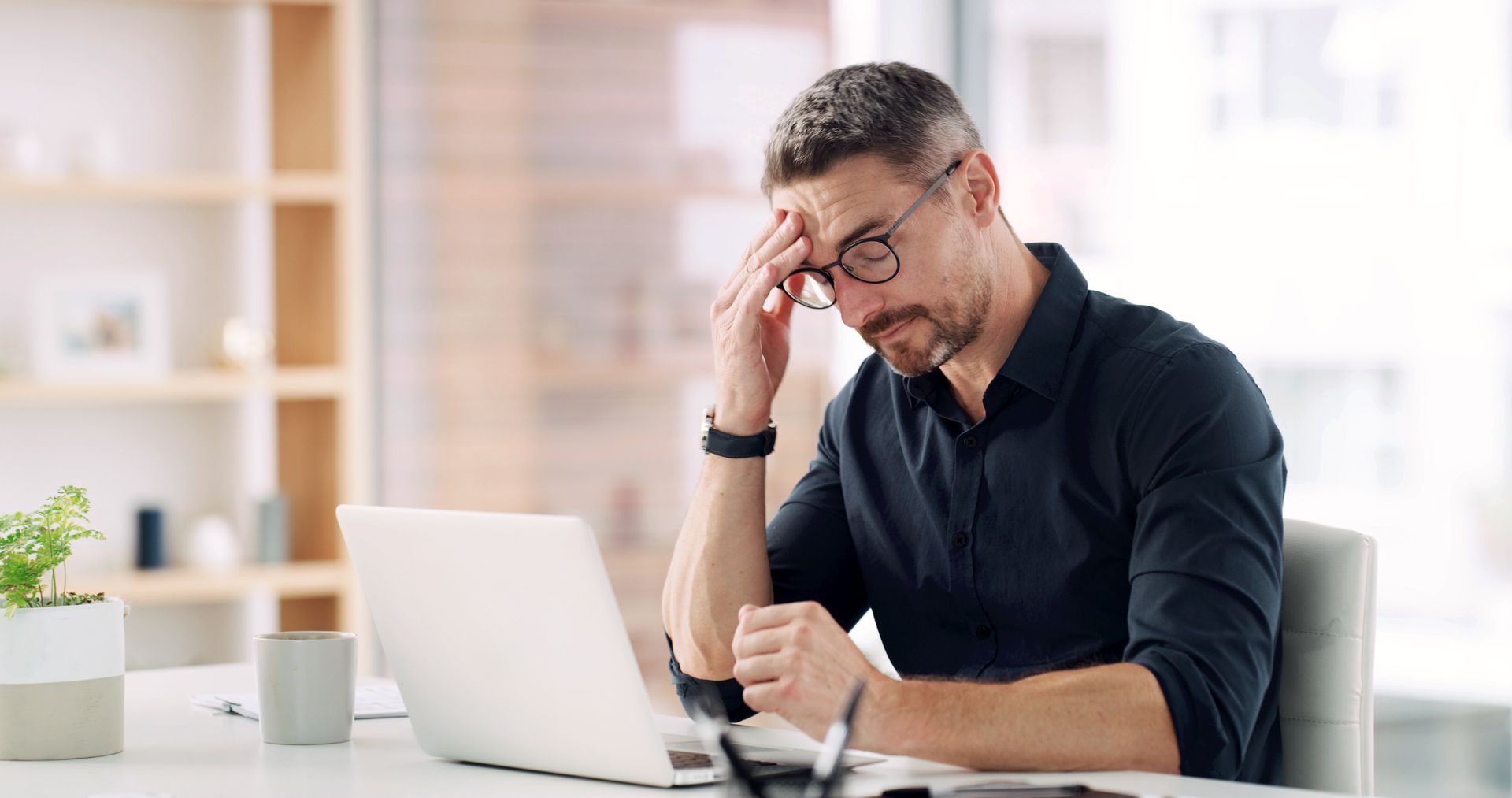 Man sitting at a desk, rubbing his forehead while working on a laptop in a bright office