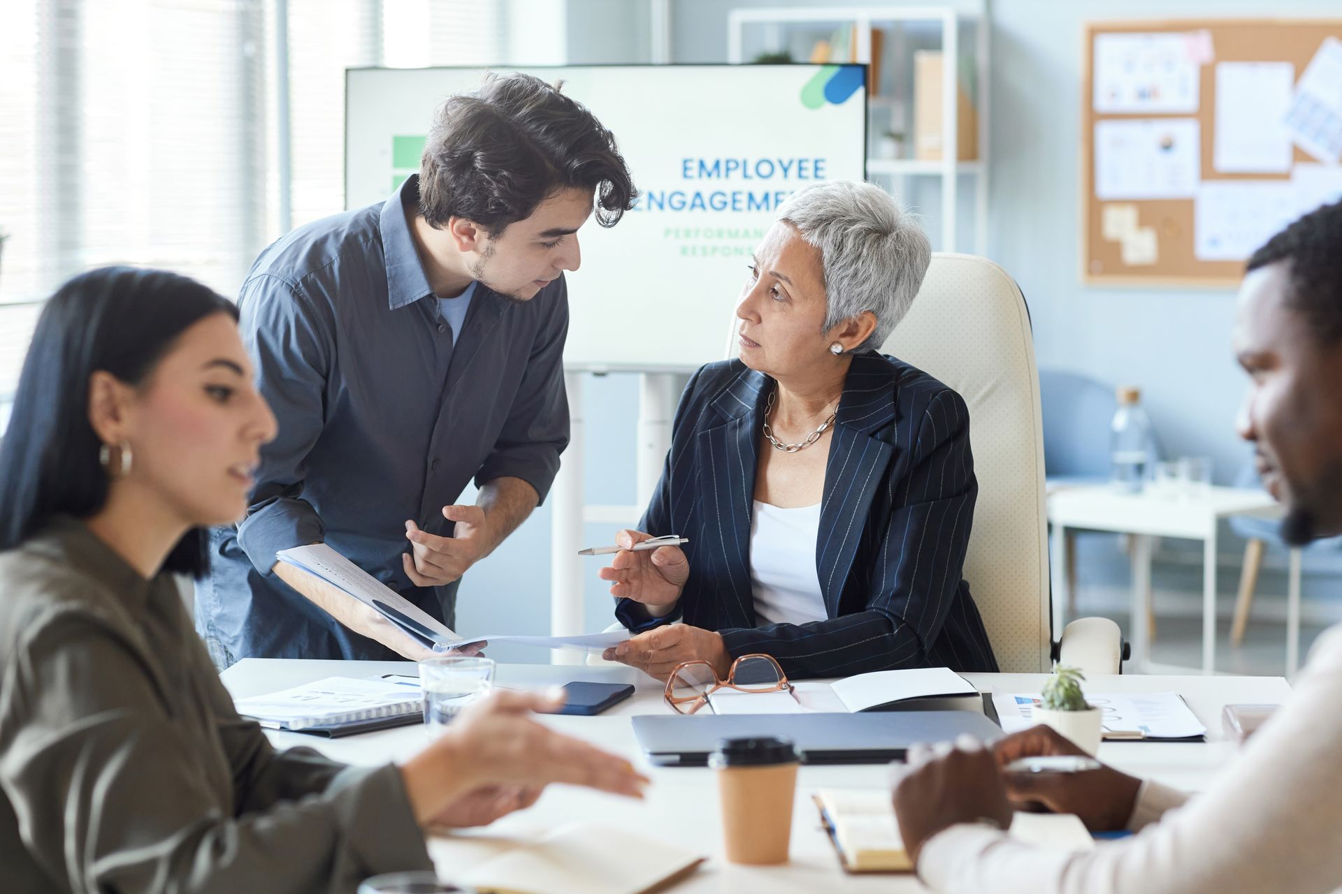 Colleagues in a meeting room discussing documents around a table, with a presentation board behind them