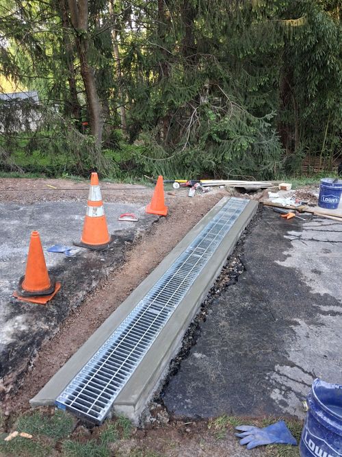 Roadwork trench with metal drainage grate, orange cones, and construction debris beside a paved path.