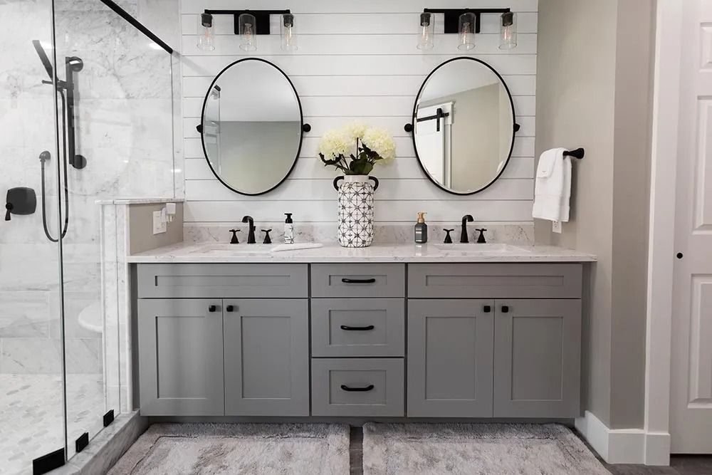 Modern double-sink bathroom vanity with gray cabinets, two round mirrors, wall sconces, and a vase of flowers.