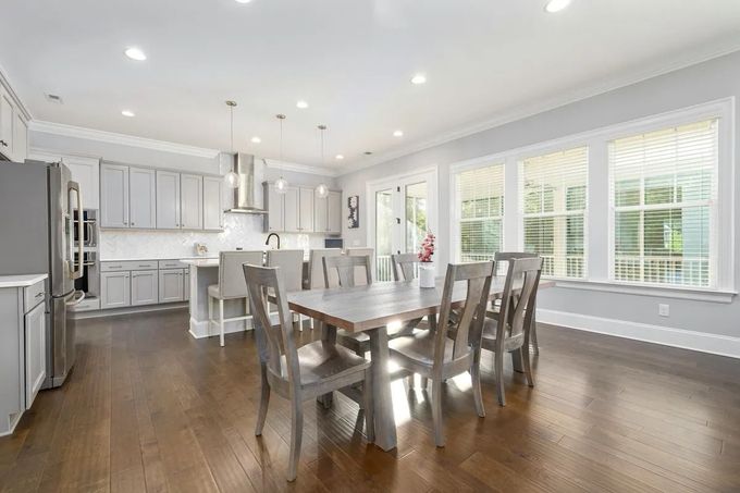 Bright modern kitchen with white cabinets, stainless appliances, island, dining table, and large windows with blinds