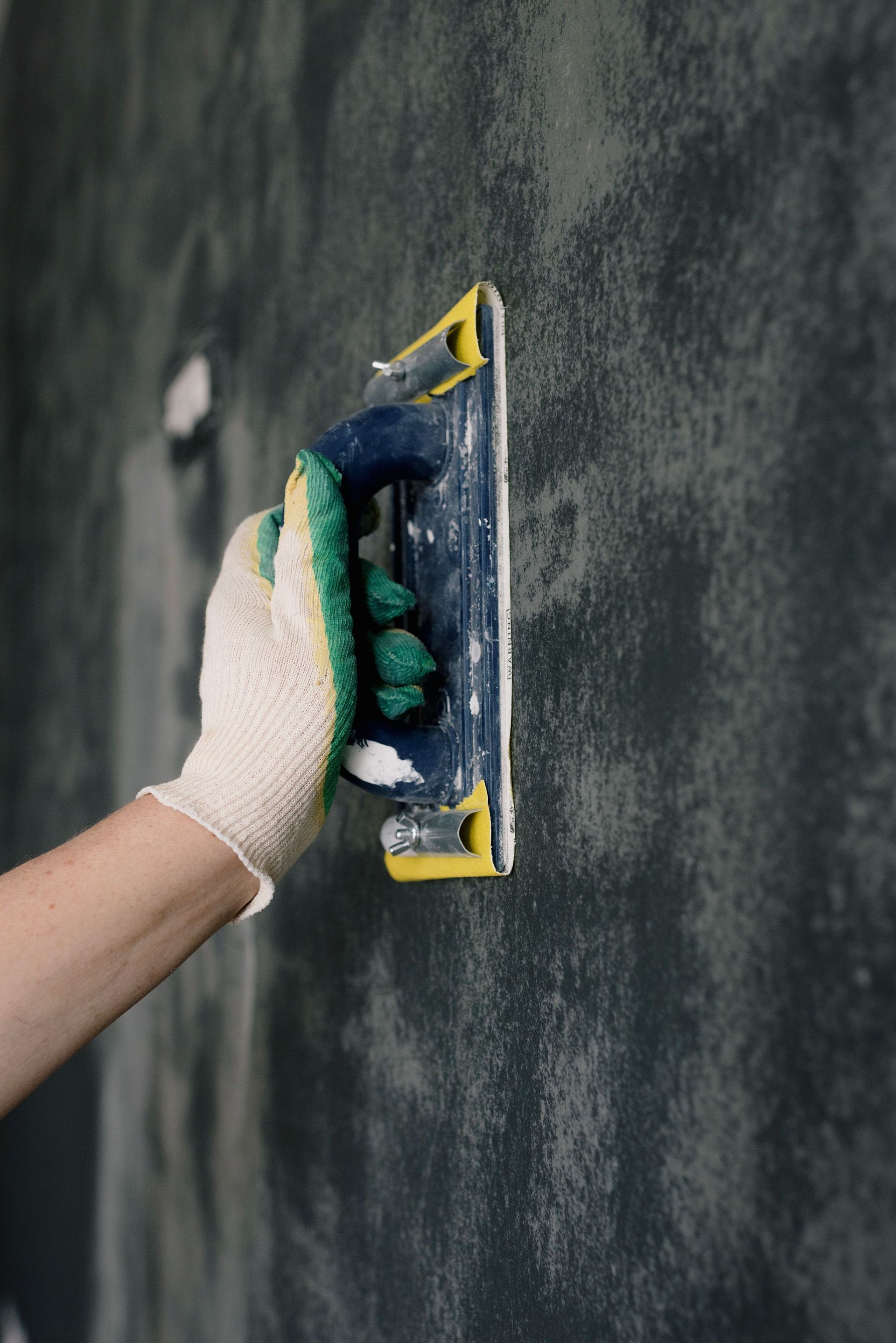 Gloved hand sanding a dark wall with a sanding block