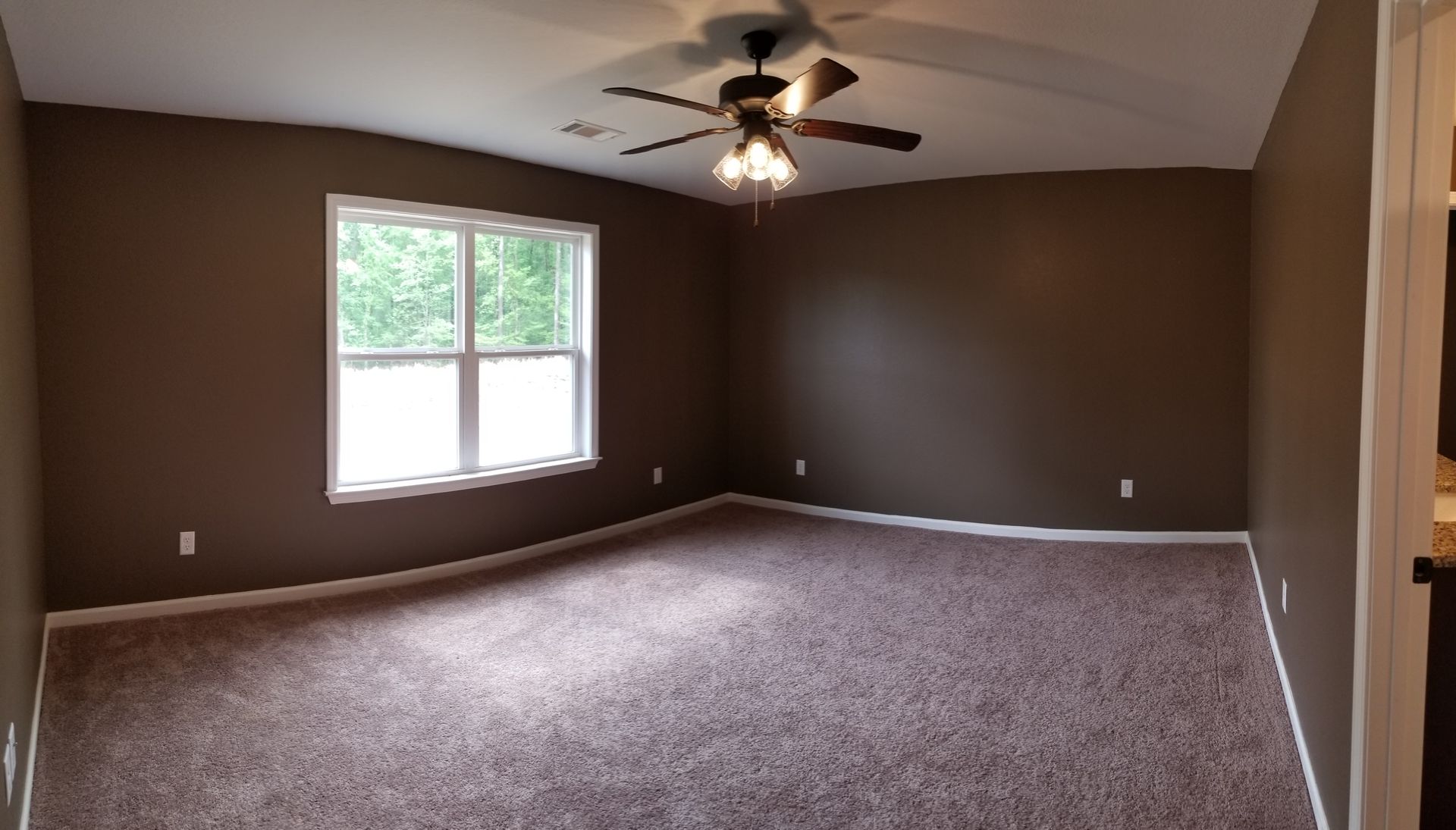Empty bedroom with brown walls, carpet, and ceiling fan. A window lets in natural light.