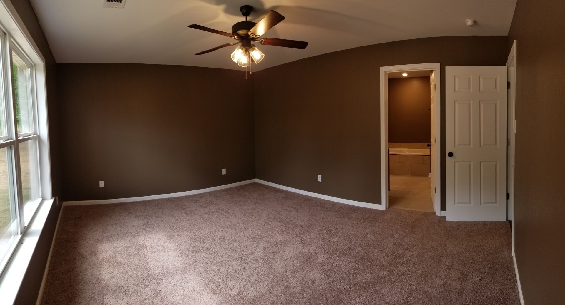 Spacious bedroom with brown walls, beige carpet, a large window, and a door leading to a bathroom.